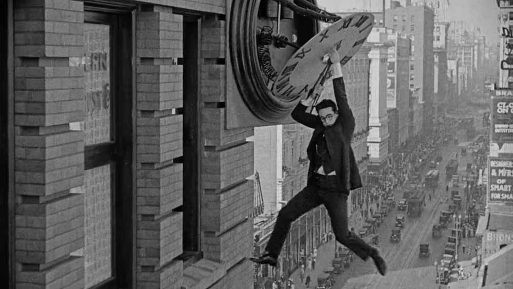 Still from Safety Last! (1923) depicting Harold Lloyd hanging precariously from the hands of a clock on the side of a building